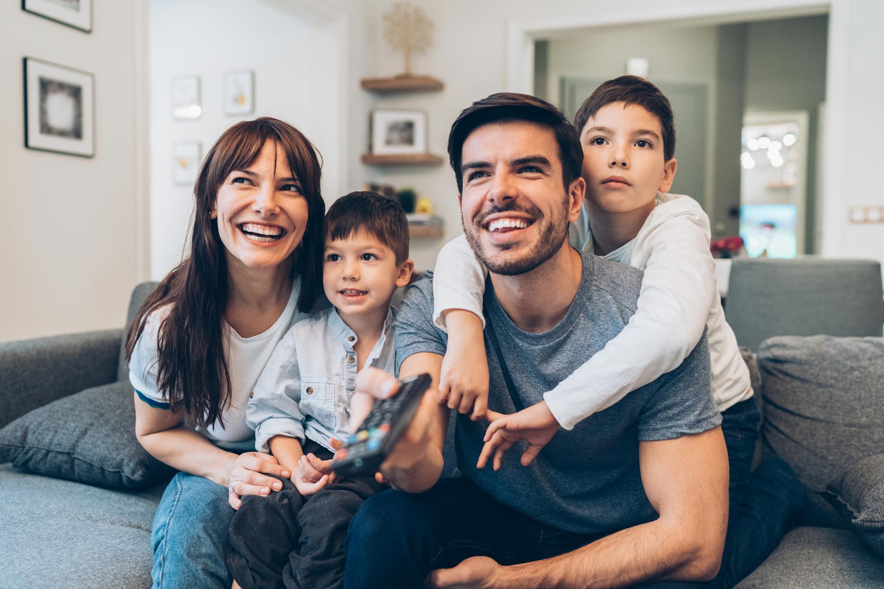 Family of four sits together on a couch in a comfortable living room, smiling while watching television and holding a remote.