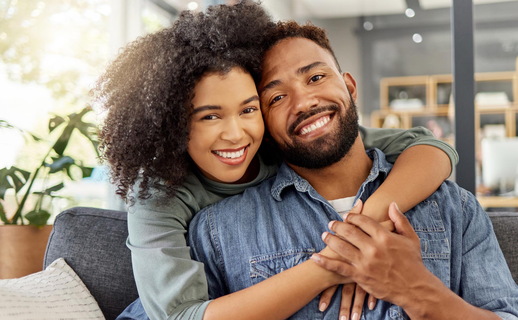 Smiling woman embraces a man from behind on a couch in a comfortable, sunlit living room